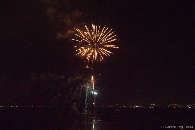 Bright fireworks burst in the night sky above Lake Suwa, with city lights visible along the distant shoreline. The reflections of the fireworks shimmer beautifully on the water’s surface.
