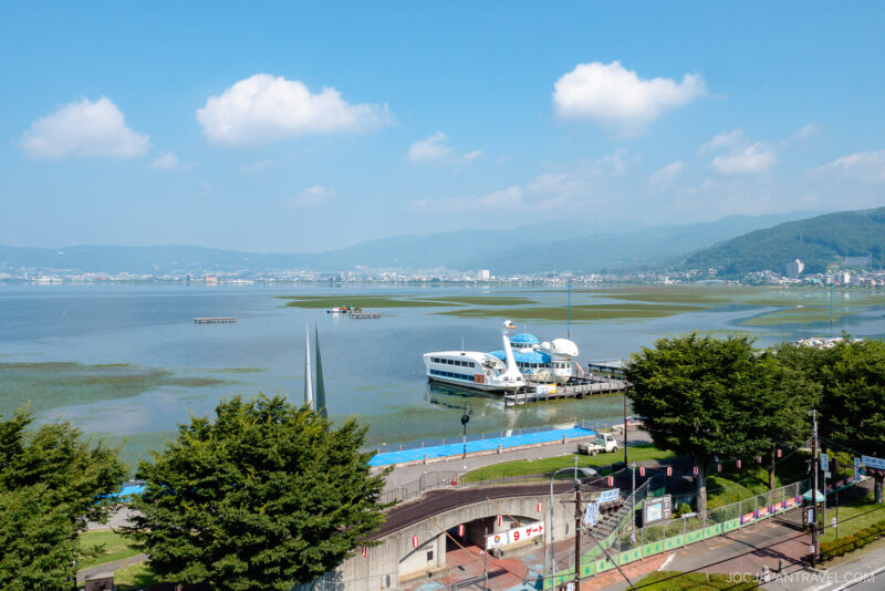A sunny Lake Suwa scene with a docked white and blue sightseeing boat, lush trees in the foreground, calm water, and distant mountains and city buildings under a blue sky with scattered clouds.