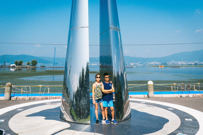 Two children stand smiling by a tall, shiny metal sculpture on the Lake Suwa waterfront promenade, with calm water, distant mountains, and a clear blue sky in the background.