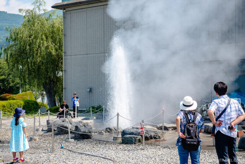 People stand around a roped-off area watching a geyser erupt with steam and water shooting into the air near a building, on a sunny day at Lake Suwa. Some wear hats and summer clothing, with trees and mountains visible in the background.