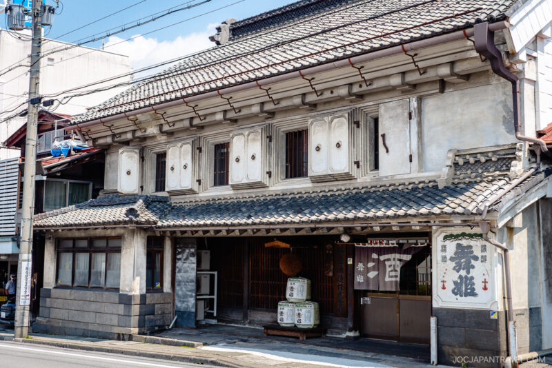 Traditional Japanese building with a tiled roof and white plaster walls, featuring wooden shutters and sake barrels outside. A fabric sign hangs at the entrance, while power lines run above the street near Lake Suwa.