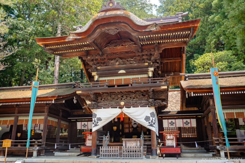 Traditional Japanese Shinto shrine near Lake Suwa, featuring an ornate wooden entrance, intricate carvings, a white curtain with black emblems, and surrounded by green trees on a sunny day.