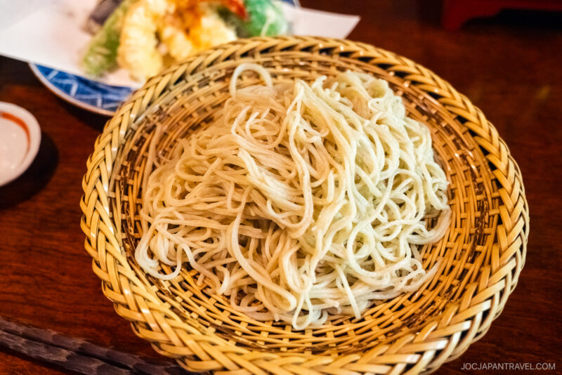 A close-up of a woven basket filled with plain soba noodles on a wooden table near Lake Suwa, with a small dish of dipping sauce and a plate of assorted tempura in the background.