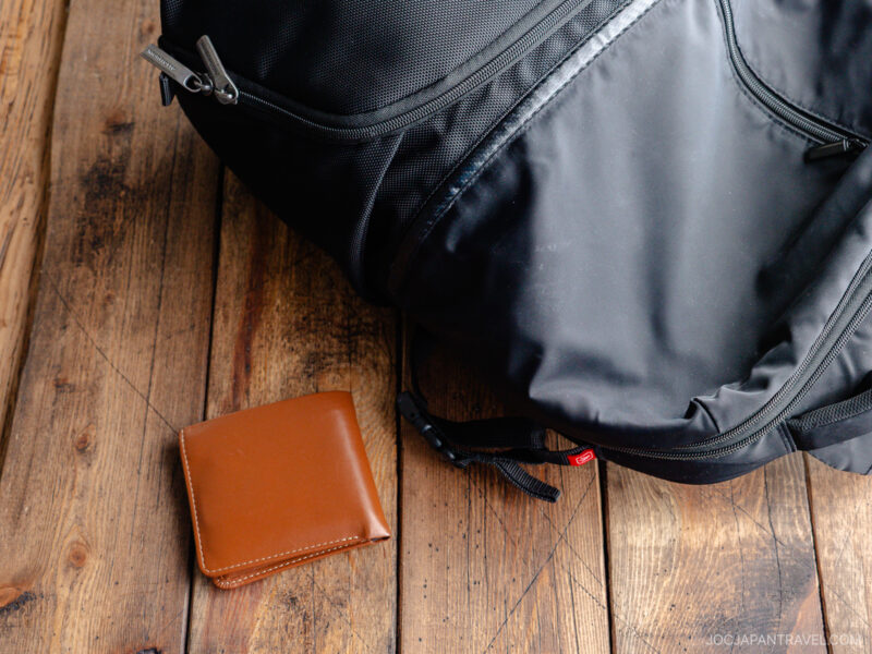 A black backpack and a closed brown leather lost wallet rest on a rustic wooden surface.