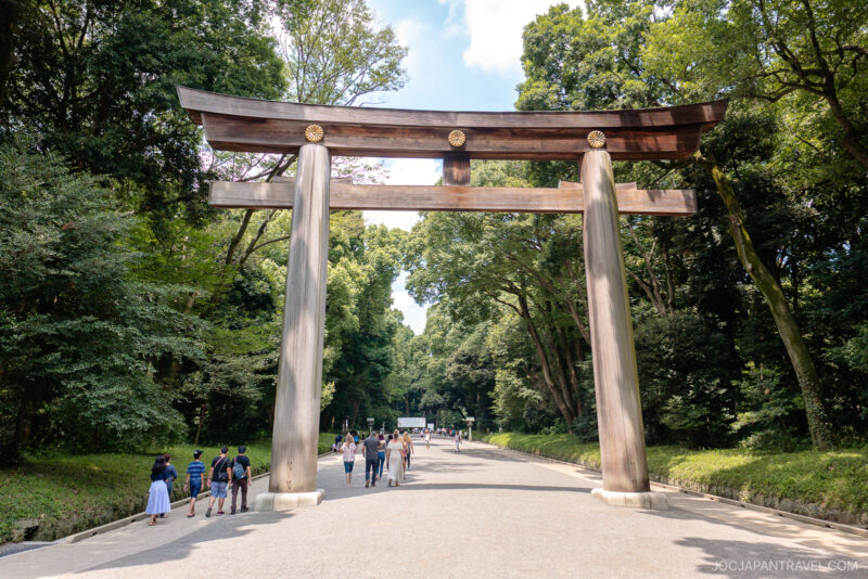 Visitors walk under a large wooden torii gate surrounded by tall green trees on a wide path at Meiji Jingu Shrine in Tokyo, Japan, on a sunny day.