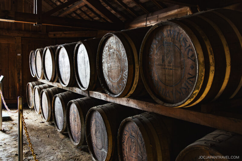 Wooden barrels are stacked in two rows inside a dimly lit, rustic storage room at the Nikka Whisky Yoichi Distillery. Some barrels have faded markings, and sunlight filters through gaps in the wooden walls and slanted roof.
