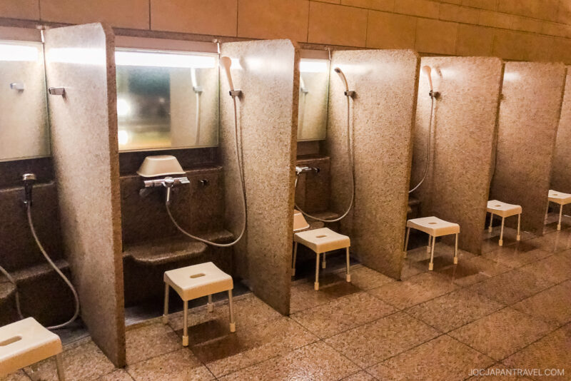 A row of partitioned shower stations in a Japanese bathhouse, each with a handheld showerhead, mirror, small stool, and washbowl—showing how to enjoy onsen by cleansing thoroughly in this clean, well-lit area with elegant stone surfaces.