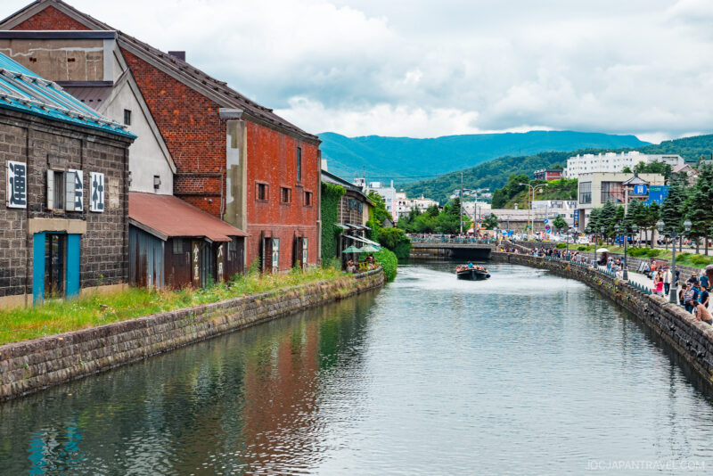 A scenic canal in Otaru lined with old warehouses and brick buildings, with people strolling along the waterfront and a boat cruising on the water, set against a backdrop of green hills and cloudy skies.