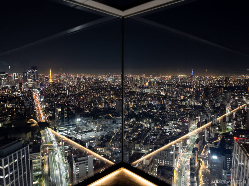 A nighttime cityscape of Tokyo viewed from a high-rise observation deck like Shibuya Sky, with bright city lights, dense buildings, and reflections on tall glass windows forming a striking V-shaped perspective.