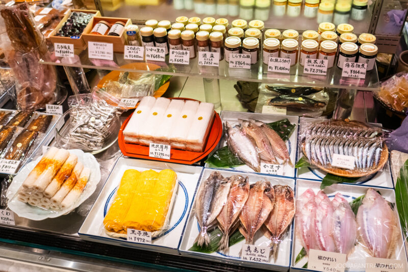 A display case in a Japanese market, reminiscent of the Isetan food floor, features packaged seafood, fish fillets, yellow tamagoyaki, jars of condiments, and labeled price tags in Japanese.