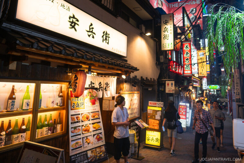 A lively alley in Japan at night, often called memory lane, features illuminated signs, restaurant menus with food and drink images, and people walking under colorful neon lights.