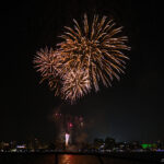 Summer Fireworks Festivals in Japan (花火大会) Bright golden fireworks burst in the night sky above a city skyline, their reflections shimmering on the water below. This festive scene resembles those in a hanabi guide, perfectly capturing the spirit of celebration.