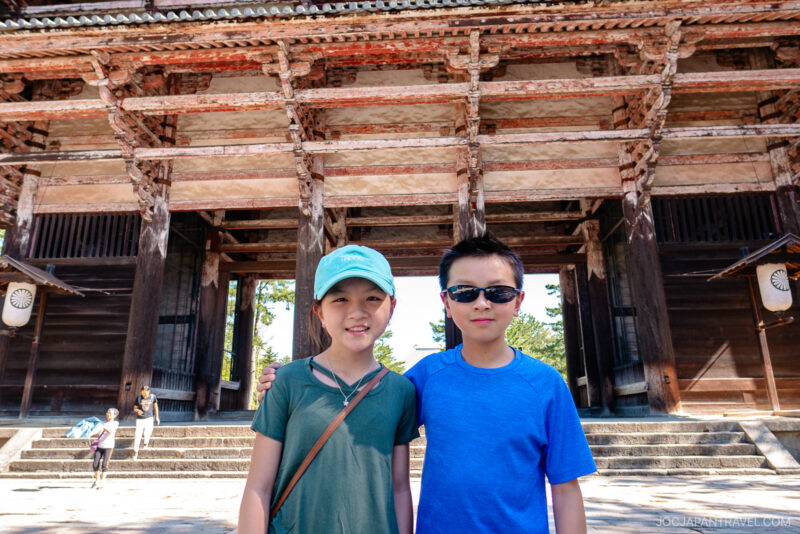 Two children stand smiling in front of the grand Todaiji wooden gate. One sports a mint cap and brown crossbody bag; the other wears sunglasses and a blue shirt. The gate features intricate woodwork and weathered paint.