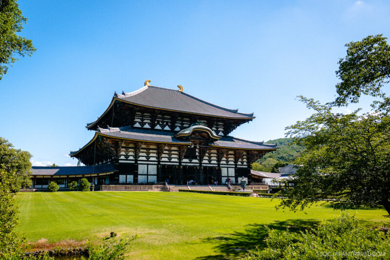 Large traditional Japanese temple, such as Todaiji, with a dark tiled roof and ornate wooden architecture, set amid a green lawn and trees under a clear blue sky.