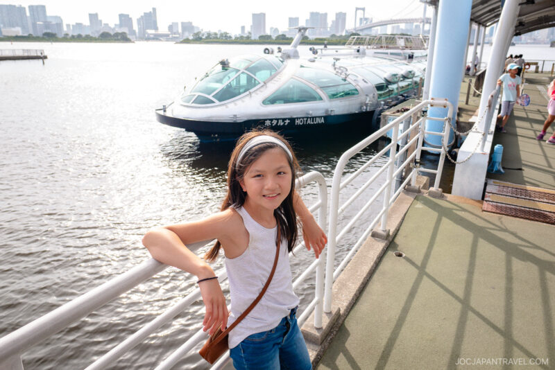 A young girl in a white tank top smiles while leaning on a railing by the waterfront, with the futuristic HOTALUNA Tokyo Cruise boat docked behind her and a city skyline in the background.