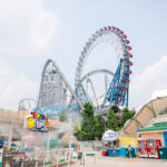 A bright, sunny day at an amusement park like Tokyo Dome City with large, looping roller coasters and a Ferris wheel. Colorful signs, information booths, and a restroom area are visible in the foreground.