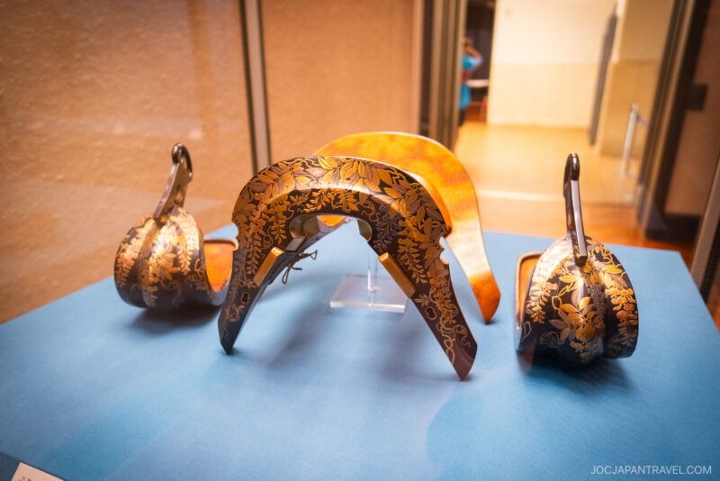A traditional Japanese saddle with ornate gold floral patterns is displayed on a blue surface at the Tokyo National Museum. The saddle and its components are elegantly crafted and polished, showcasing exquisite artistry in a museum setting.