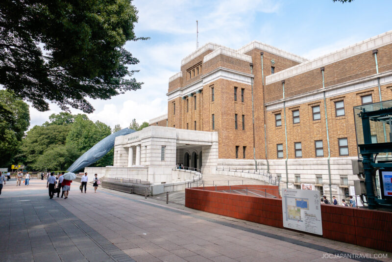 People walk near the main entrance of the Tokyo National Museum of Nature and Science, a large historic brick building with white stone features. A life-sized blue whale statue stands on the left, while trees border the walkway.