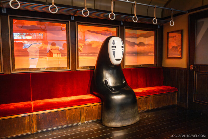 A life-sized statue of No-Face from Spirited Away sits alone on a red velvet train seat in a studio set resembling a train carriage, reminiscent of the iconic scene recreated at Ghibli Park, with sunset scenery visible through the windows.