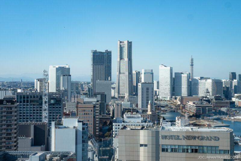 A cityscape view of Yokohama, Japan, featuring modern high-rise buildings under a clear blue sky. The prominent Hotel New Grand and the iconic Yokohama Marine Tower are visible in the foreground.
