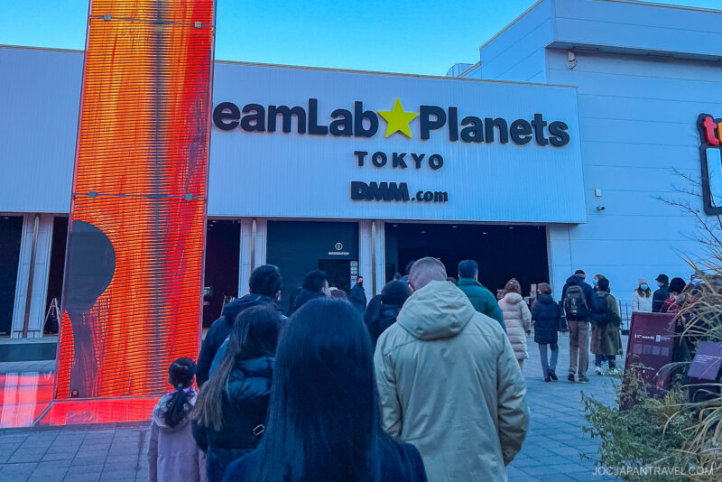 A group of people stand in line outside the entrance to teamLab Planets Tokyo, a digital art museum with a large sign above the doors and a tall, illuminated red pillar on the left.
