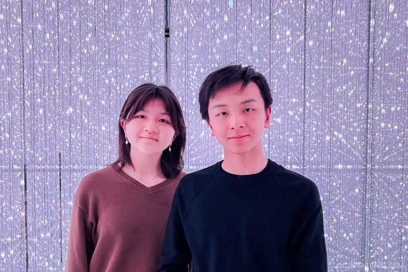 Two young people stand side by side, smiling at the camera in front of a sparkling background filled with hanging lights, capturing the bright and festive atmosphere of teamLab Planets Tokyo.