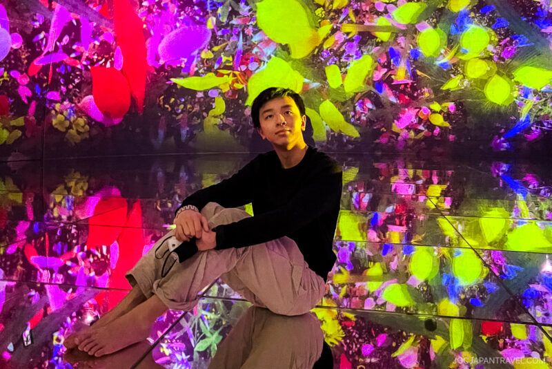 A young person sits barefoot on a reflective floor at teamLab Planets Tokyo, surrounded by vivid, colorful digital art projections of flowers and leaves, creating a mesmerizing, immersive effect.