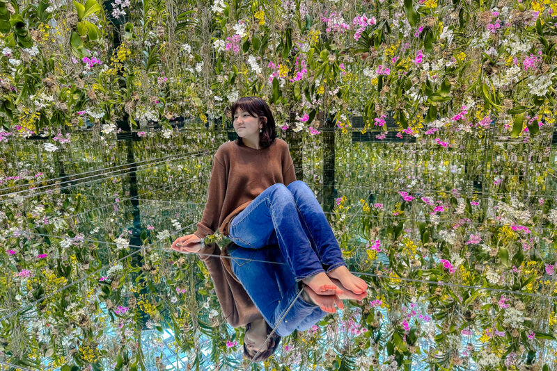 A woman sits barefoot on a reflective floor at teamLab Planets Tokyo, surrounded by hanging flowers and green plants, creating a mirrored, immersive garden effect. She wears a brown sweater and blue jeans, appearing calm and contemplative.