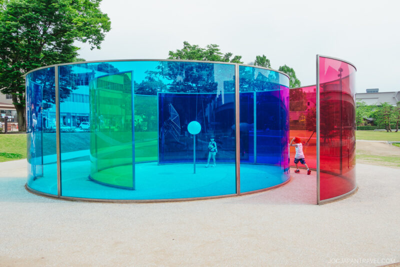 A circular outdoor art installation at the 21st Century Museum of Contemporary Art features tall, curved glass panels in blue, yellow, and red. Children play inside as colorful light and reflections dance on the ground, framed by trees and buildings in the background.