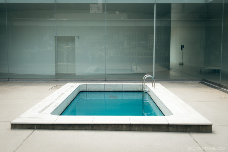 A rectangular art installation resembling a swimming pool with clear blue water, set in a minimalist courtyard surrounded by glass walls and a concrete floor at the 21st Century Museum of Contemporary Art.