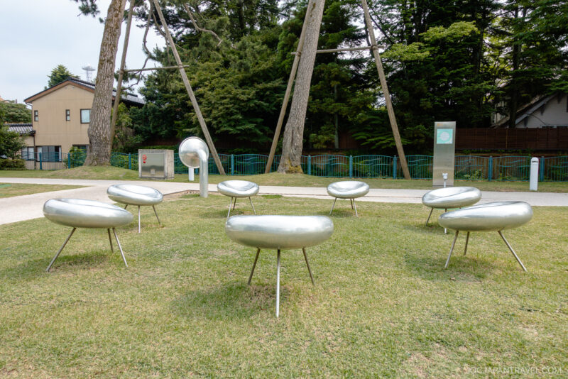 Seven metallic, circular sculptures with three legs each are arranged in a circle on a grassy area in a park near the 21st Century Museum of Contemporary Art, with trees and houses in the background.
