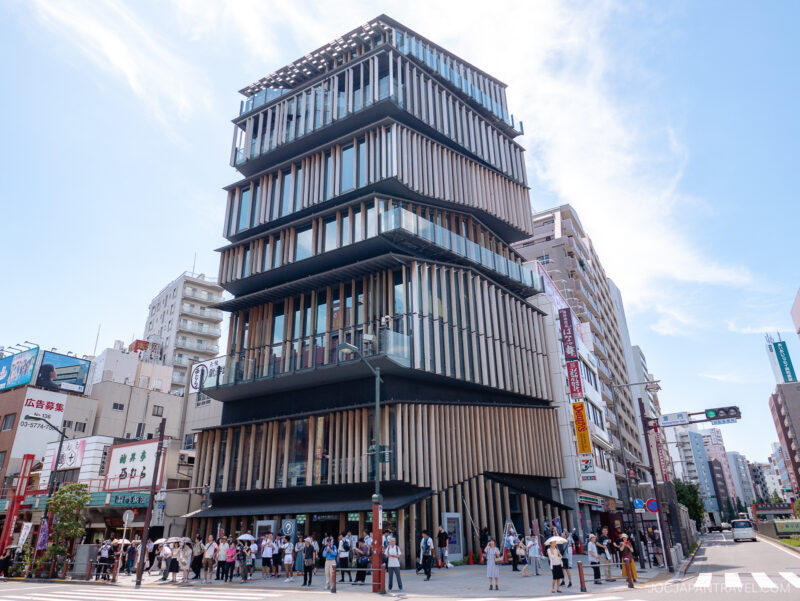 A modern multi-story building with staggered floors and vertical wooden slats stands on a busy city street, surrounded by smaller buildings, with many people gathered in front under a partly cloudy sky.