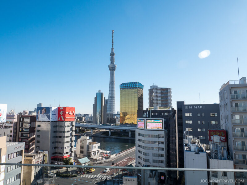 View of Tokyo cityscape with the Tokyo Skytree tower centered, surrounded by modern buildings, a river, and clear blue sky in the background.