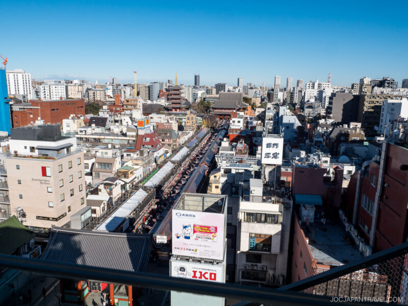 Aerial view of Tokyo’s Asakusa district, showing a busy shopping street lined with shops, with the Senso-ji Temple in the background and modern city buildings under a clear blue sky.