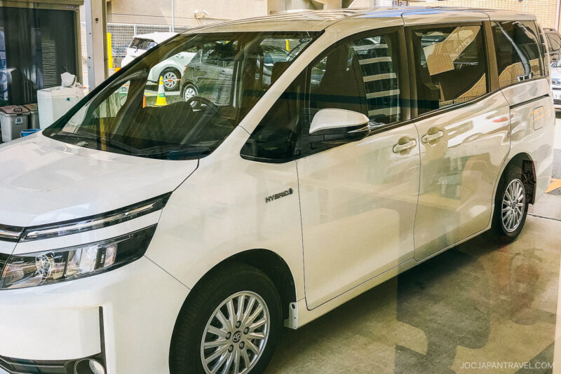 A white HYBRID minivan, highlighted in this Japan driving guide, is parked indoors near a garage or service area, with reflections visible on its windows and body.
