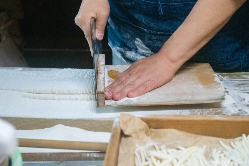 A person uses a large knife to cut dough into strips on a floured wooden board, preparing homemade noodles—a scene reminiscent of traditional food in Takayama.