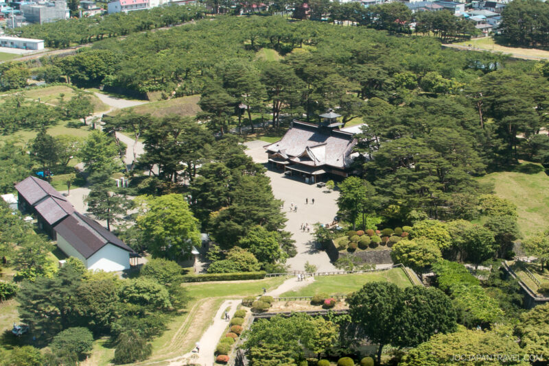 Aerial view of Fort Goryokaku, a historic Japanese building surrounded by dense green trees and gardens, with walkways and people scattered around the complex. Smaller white buildings are nearby, and a city is visible in the background.