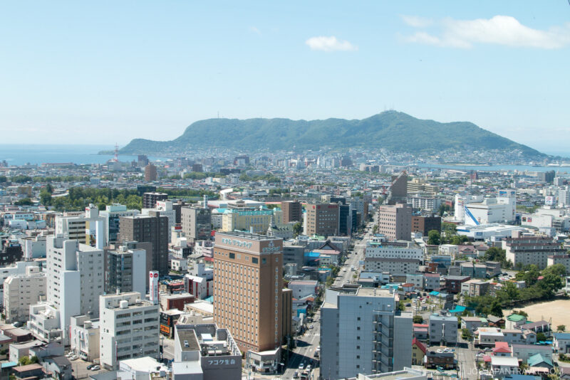 A cityscape with numerous mid-rise buildings in the foreground and the historic Fort Goryokaku nestled against a lush green mountain in the background under a clear blue sky.