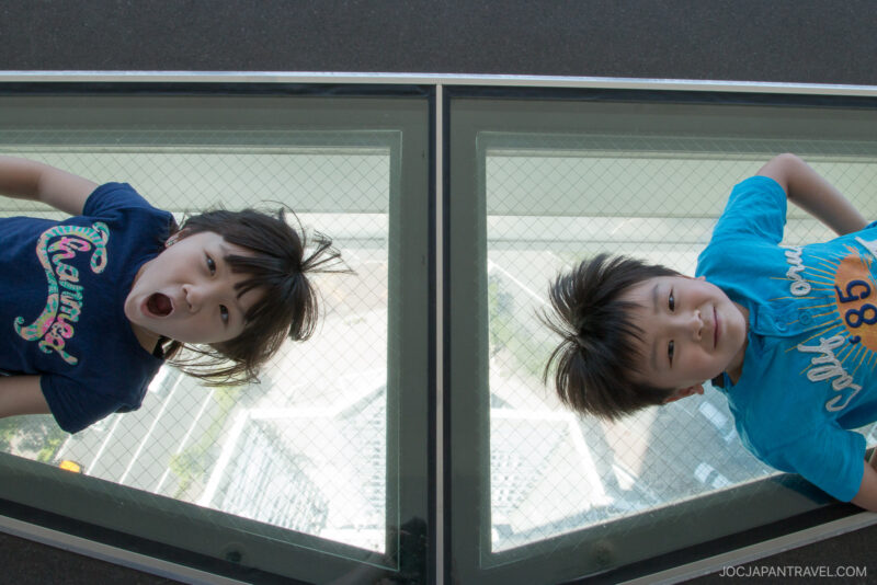 Two children lie on a glass floor panel at Fort Goryokaku, looking up. One child has an open-mouthed expression of surprise, while the other smiles. The ground and a metal frame are visible through the glass beneath them.