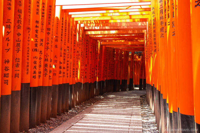 A pathway lined with rows of vibrant orange torii gates featuring black Japanese inscriptions creates a tunnel effect at Fushimi Inari Shrine in Kyoto, Japan. Sunlight filters through the gates, casting shifting shadows on the path.