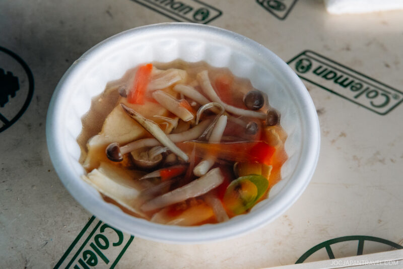 A white bowl filled with clear soup featuring mushrooms, tofu slices, bean sprouts, and vegetables sits on a surface with green text and hachinohe logos.