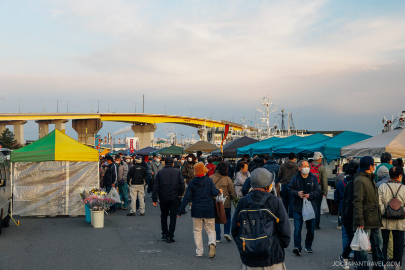 People walk through Hachinohe’s outdoor market with colorful tents and stalls, some carrying bags and wearing masks. A yellow bridge and docked boats are visible in the background under a cloudy sky.