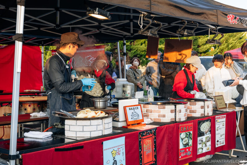 Street food vendors in Hachinohe prepare and serve hot dishes at an outdoor market stall, while customers watch and wait. The scene is lively, with steam rising from the cooking area under a black canopy.