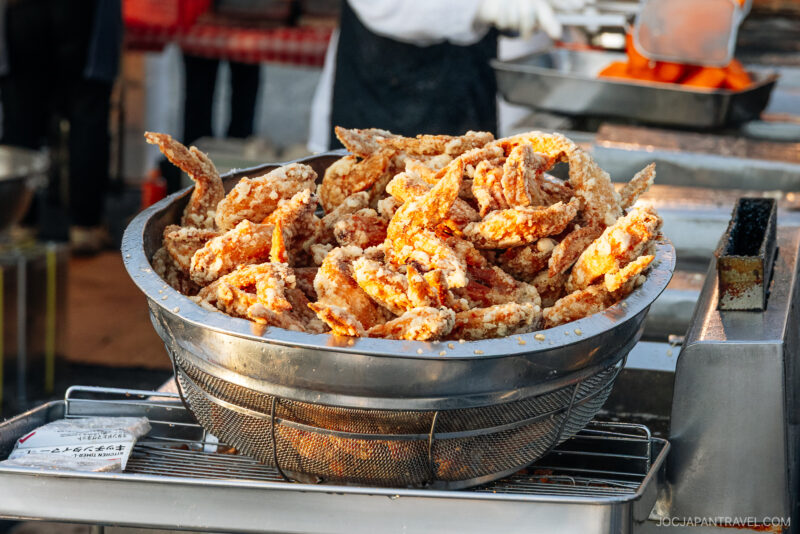 A large metal strainer filled with freshly fried chicken wings sits on a grill at a bustling Hachinohe outdoor food stall. A person in gloves prepares more food in the background.