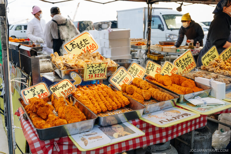 A market stall in Hachinohe displays trays of deep-fried foods with handwritten yellow signs showing prices. Vendors in masks and hats serve customers in the background, and vehicles are visible behind the stall.