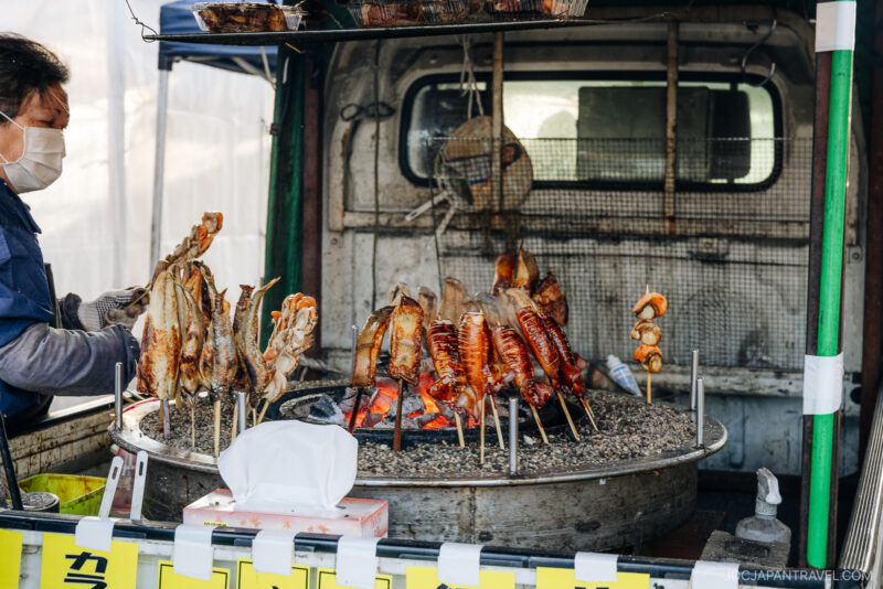A street vendor in Hachinohe wearing a mask grills whole skewered fish over a charcoal fire on a large round grill set up on the back of a truck. Grilled fish are arranged in a circle above hot coals.