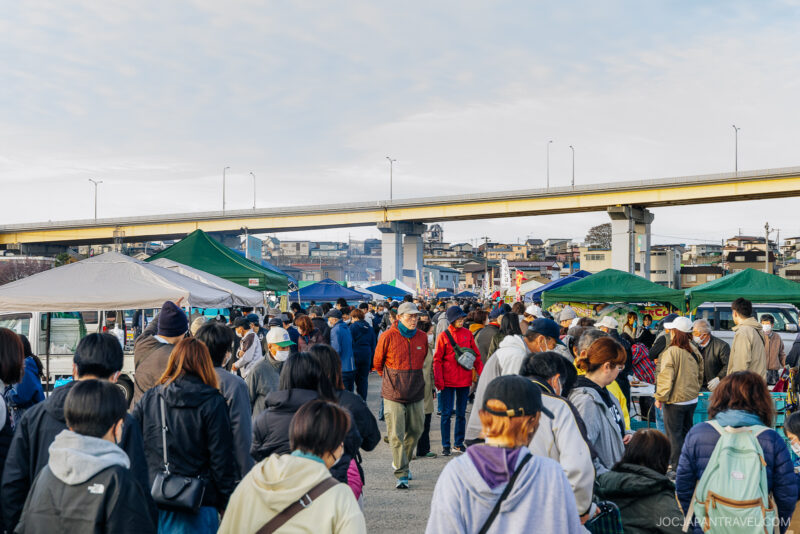 A busy outdoor market in Hachinohe with many people walking among stalls and tents, some wearing masks, under an overpass with buildings in the background and a partly cloudy sky above.