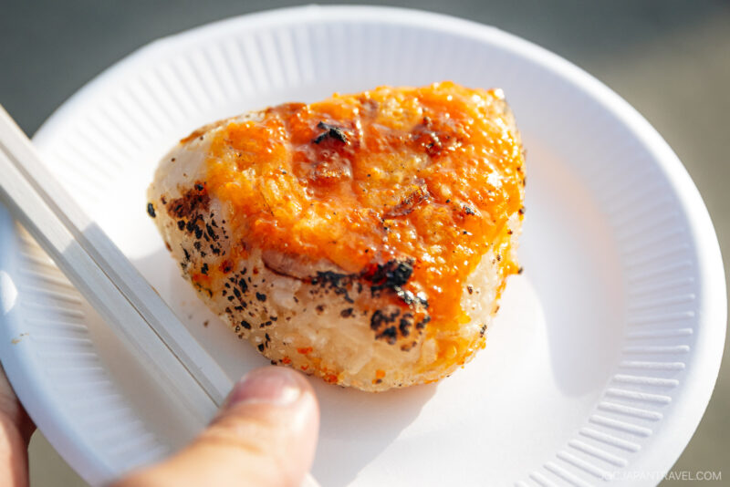 A hand holding a white paper plate with a grilled onigiri rice ball topped with a golden, crispy sauce—a delicious taste of Hachinohe—served with a pair of chopsticks on the side.