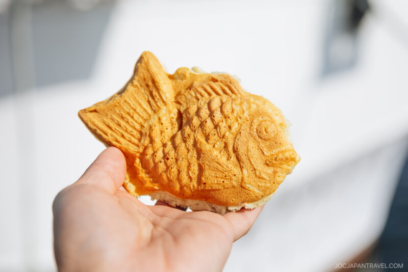 A hand holds a golden-brown, fish-shaped pastry called taiyaki, its detailed scales and fins on display. This tasty treat is popular in places like Hachinohe, with a blurred white background highlighting its artistry.