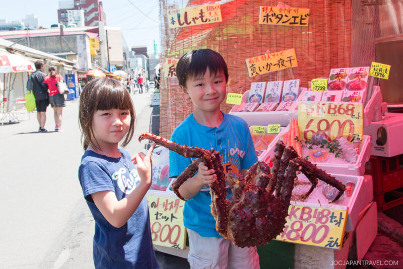 Two young children stand outdoors in front of the Hakodate Morning Market, smiling and holding up a large crab. Various seafood items with Japanese labels and prices are displayed on tables behind them.